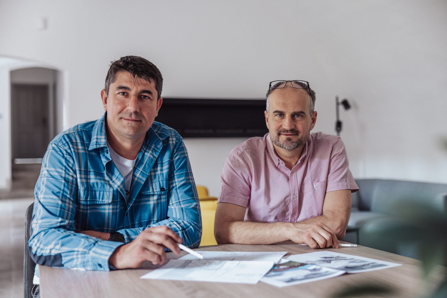 dvaja muži v strednom veku sedia pri stole s architektonickými papiermi